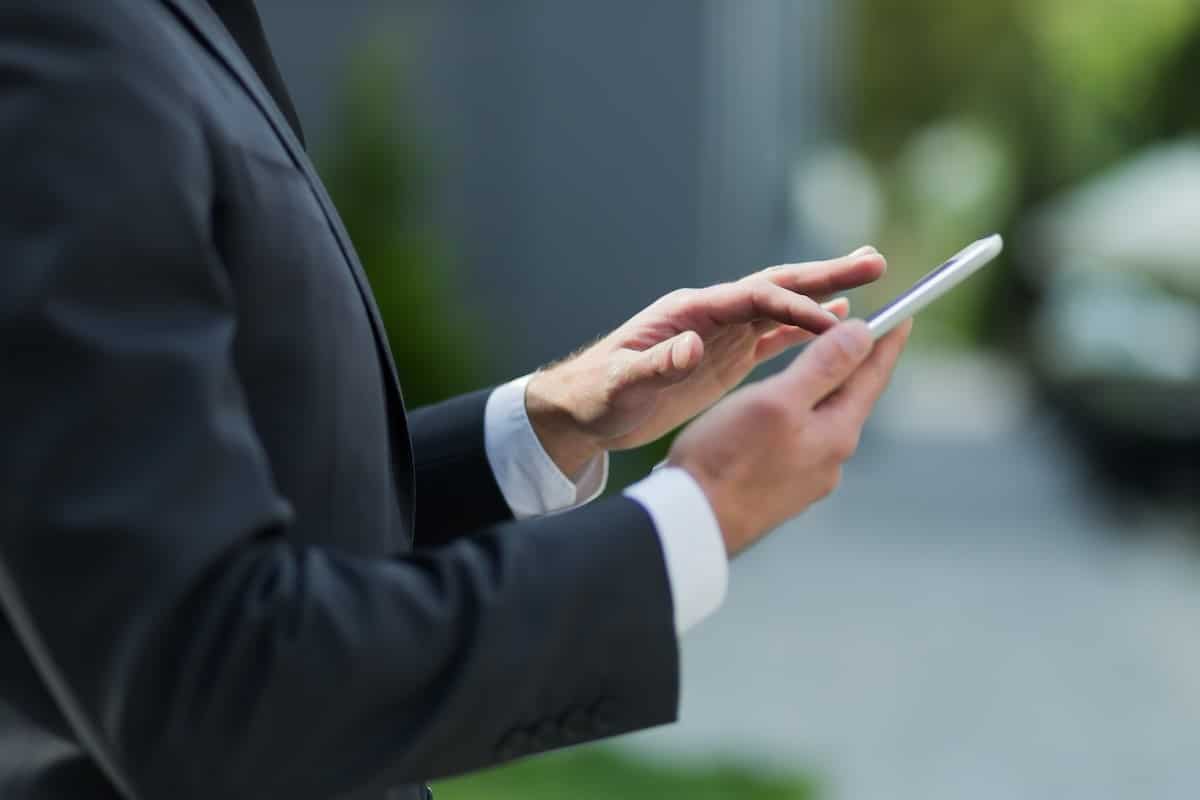 Un professionnel en costume noir consulte une tablette tactile avec les mains, dans un décor flou aux tons verts et gris, lié à la signature électronique.