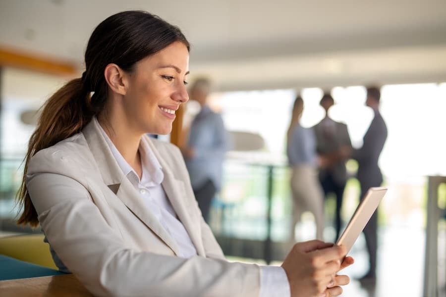 Une cadre souriante consulte une tablette numérique dans un bureau lumineux, tandis que plusieurs collègues restent flous en arrière-plan.