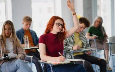 Dans une salle de classe, une étudiante rousse lève la main pendant que plusieurs camarades prennent des notes au milieu d’un cours universitaire.