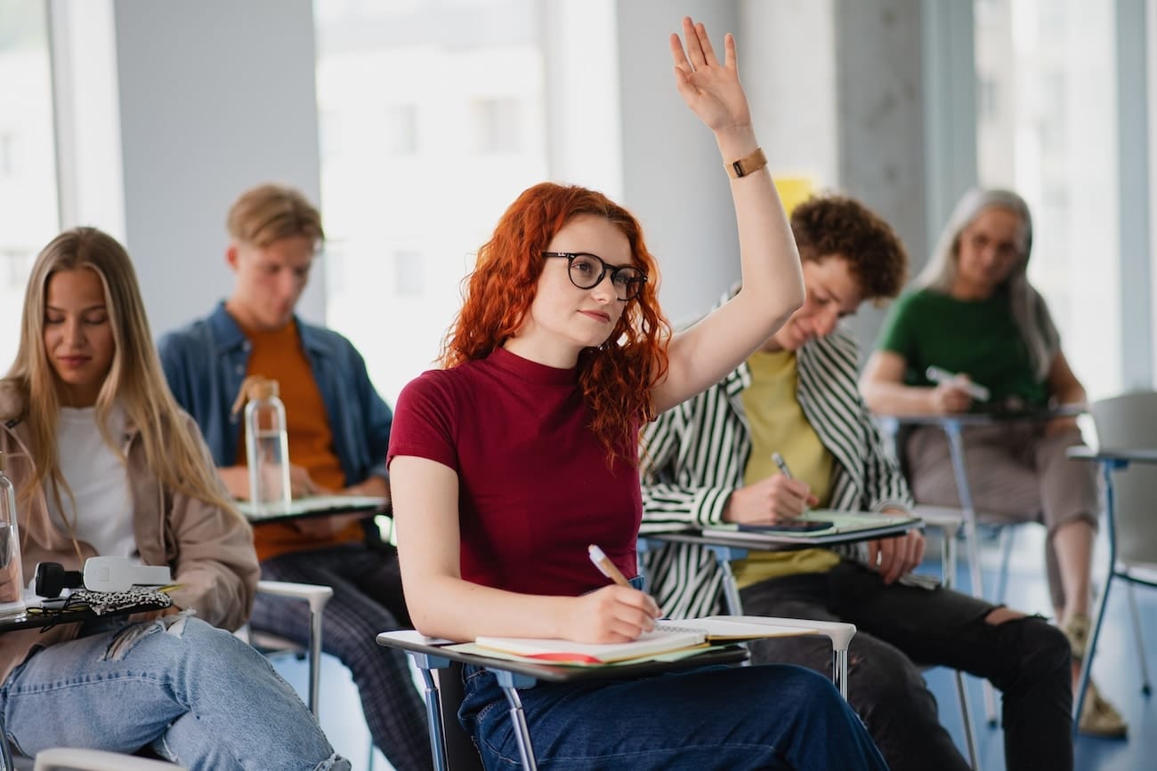 Dans une salle de classe, une étudiante rousse lève la main pendant que plusieurs camarades prennent des notes au milieu d’un cours universitaire.