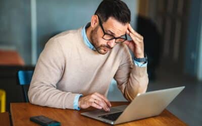 Un homme en pull beige consulte un ordinateur portable, le visage appuyé sur la main, dans une attitude de doute et de tension.