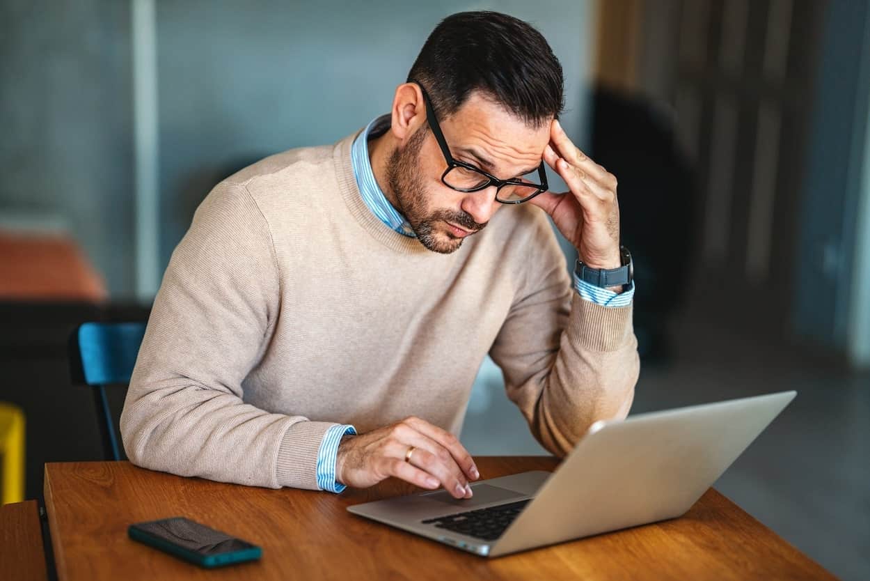 Un homme en pull beige consulte un ordinateur portable, le visage appuyé sur la main, dans une attitude de doute et de tension.