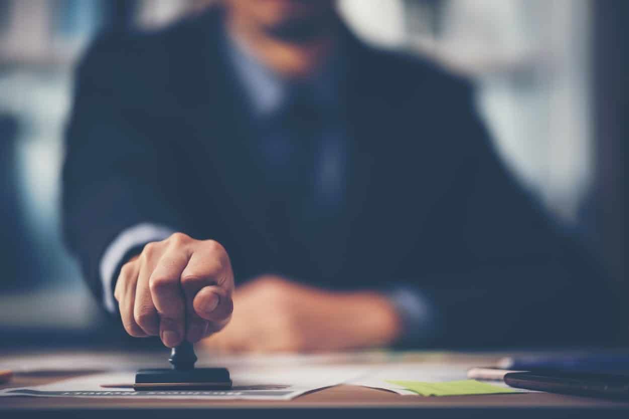 Une main en gros plan appose un cachet sur un document de bureau, avec un costume bleu foncé et un arrière-plan flou.
