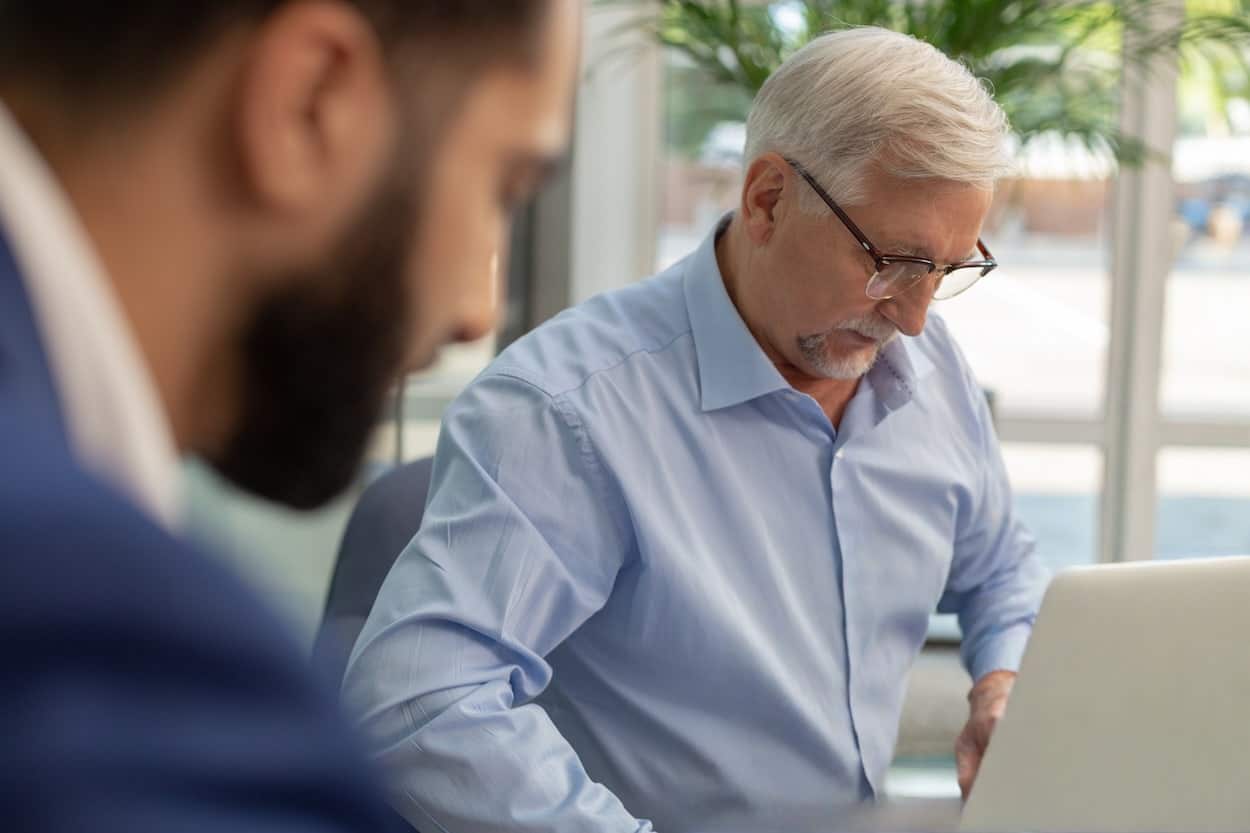 Dans un bureau lumineux, un homme âgé aux cheveux blancs consulte un ordinateur portable tandis qu’un collègue flou reste au premier plan.