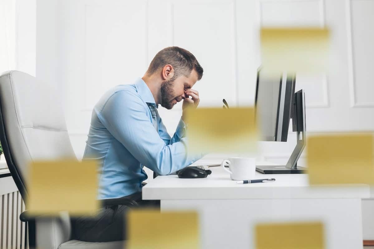 Un homme d'affaires fatigué se tient à son bureau lumineux, le front appuyé contre la main, devant un écran et des notes jaunes dispersées.
